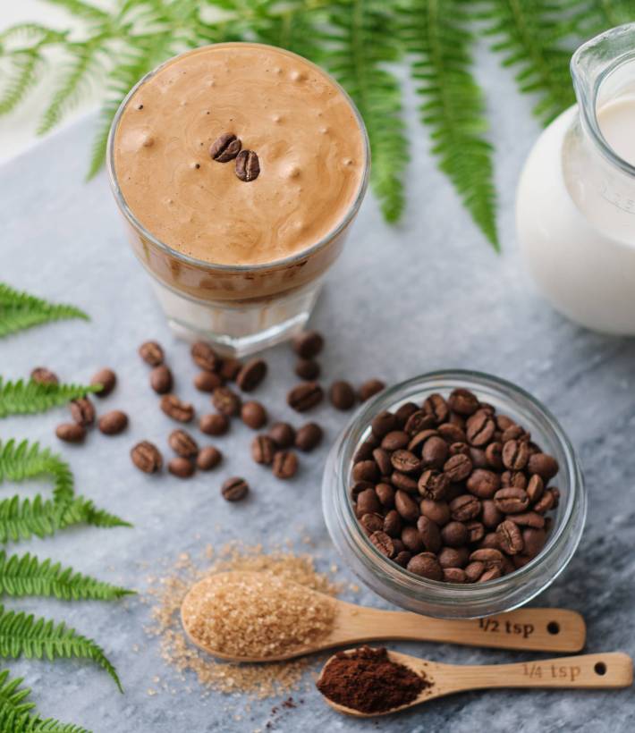 Top view of  dalgona coffee with almond milk and brown sugar, coffee beans and green plant on marble board. A glass of trendy fluffy Korean drink with whipped cream.